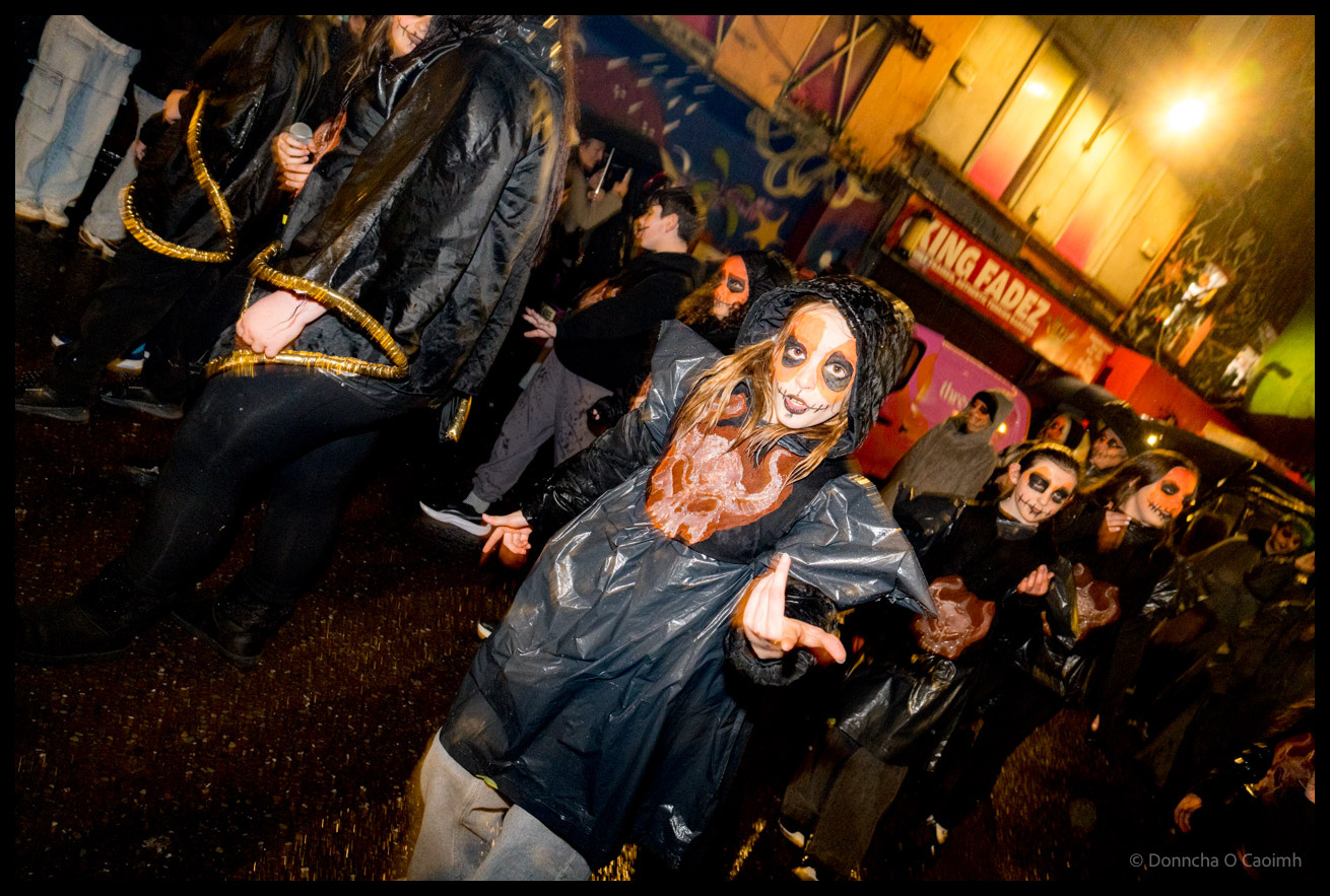 Young participant in black costume with detailed skull face paint and orange fabric details crouching dramatically during Dragon of Shandon parade on North Main Street Cork with crowd and King Fadez shop visible in background.