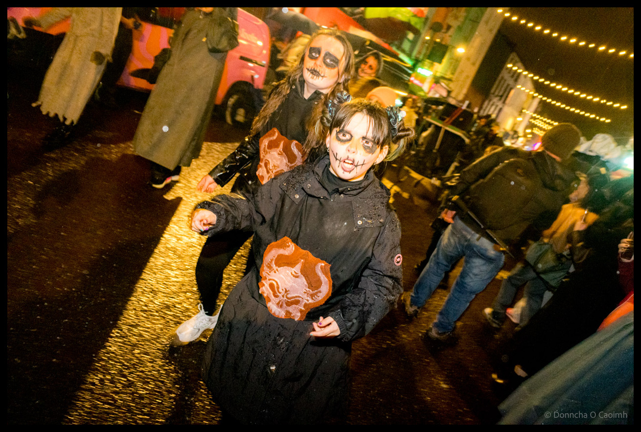 Two young participants in black clothing with skeleton face paint and orange prop masks performing energetically during Dragon of Shandon parade on North Main Street Cork at night with string lights visible overhead.