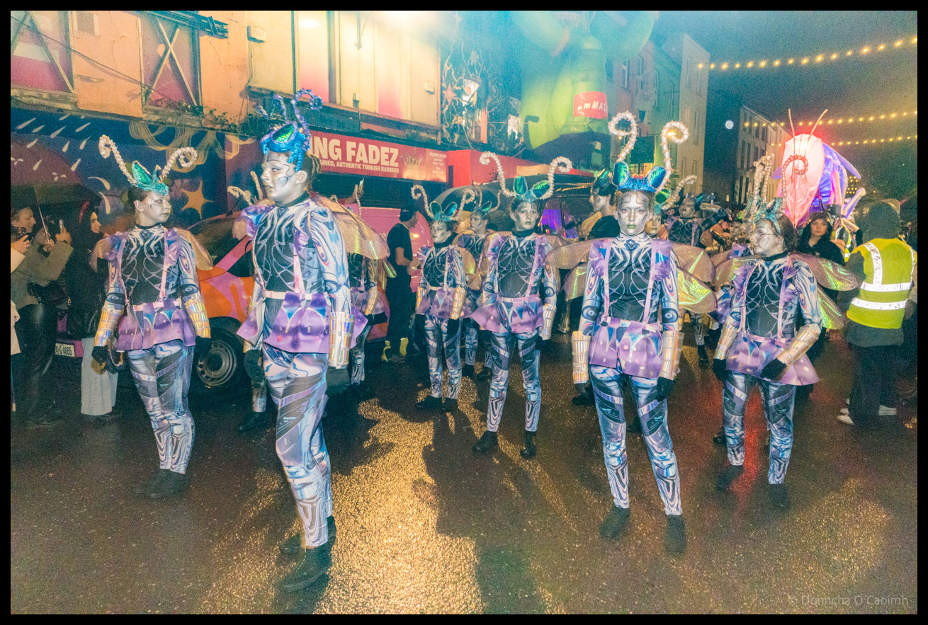 A group of dancers from Joan Denise Moriarty School of Dance in coordinated purple and silver costumes with elaborate headdresses performing choreographed routine during Dragon of Shandon parade on North Main Street, Cork, at night, with a pink and orange striped van and King Fadez shop visible in background.