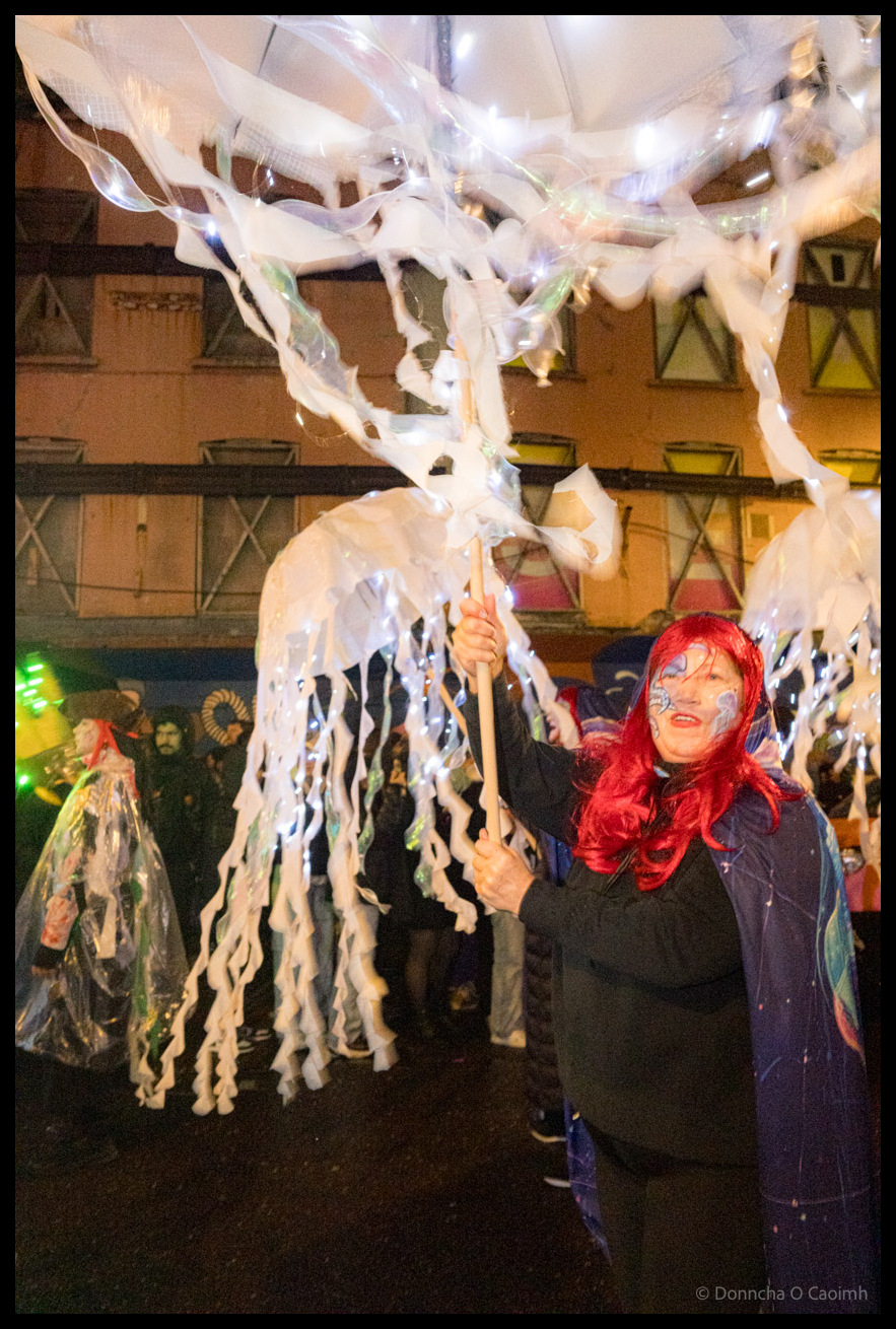 Performer with red hair and theatrical face paint in dark blue costume holds large illuminated white jellyfish puppet overhead during Dragon of Shandon parade on North Main Street, Cork, with motion blur effects and colourful building backdrop.