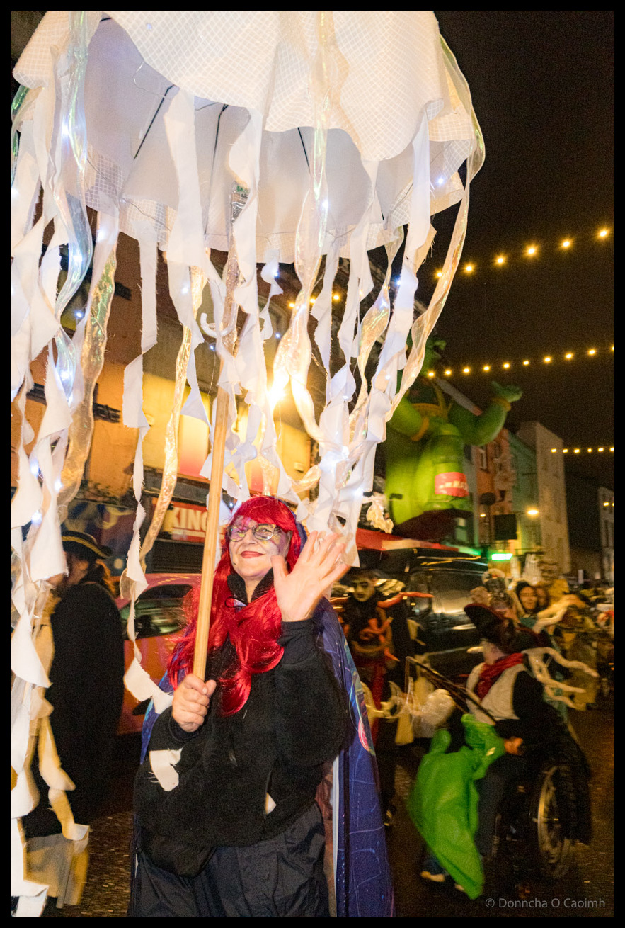 A young participant with red hair and glasses in black costume with blue cape smiles while holding a massive white jellyfish puppet with streaming tentacles during Dragon of Shandon parade on North Main Street, Cork, at night with string lights overhead.