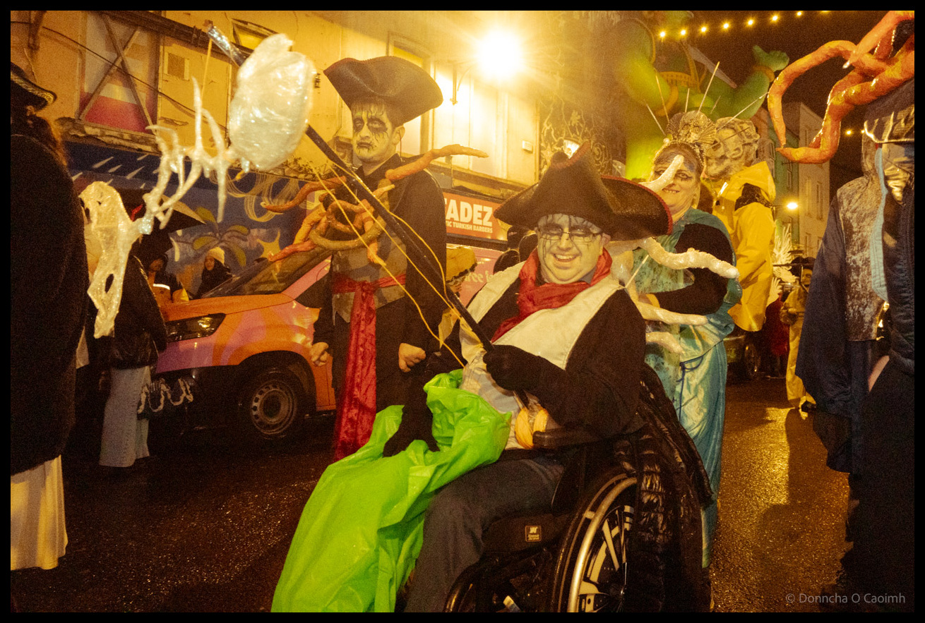 A group of costumed Dragon of Shandon parade participants, including a wheelchair user in pirate costume with white jellyfish puppet, person in red scarf, and others in theatrical makeup gathered on North Main Street, Cork, with the King Fadez shop visible in background.