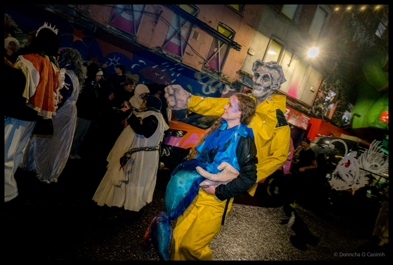 Dragon of Shandon parade participant in bright yellow raincoat carrying person in blue costume through North Main Street, Cork, at night, surrounded by Victorian-costumed performers and elaborate puppet creatures with colourful murals visible on buildings behind.