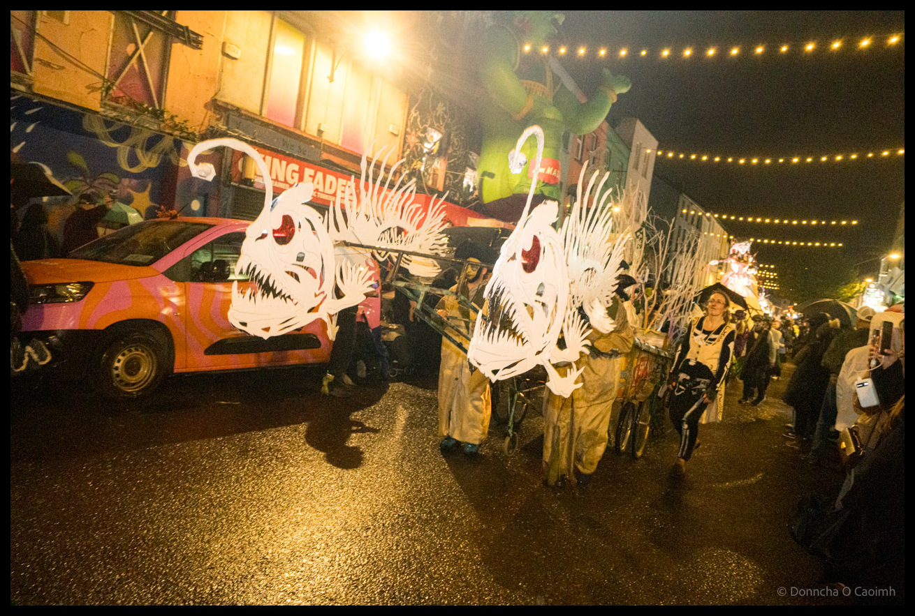 Two participants pulling a carriage with large white skeletal fish or sea creature puppets with illuminated spines parade down North Main Street, Cork, during Dragon of Shandon, with a pink and orange striped van and King Fadez shop visible in background under string lights.