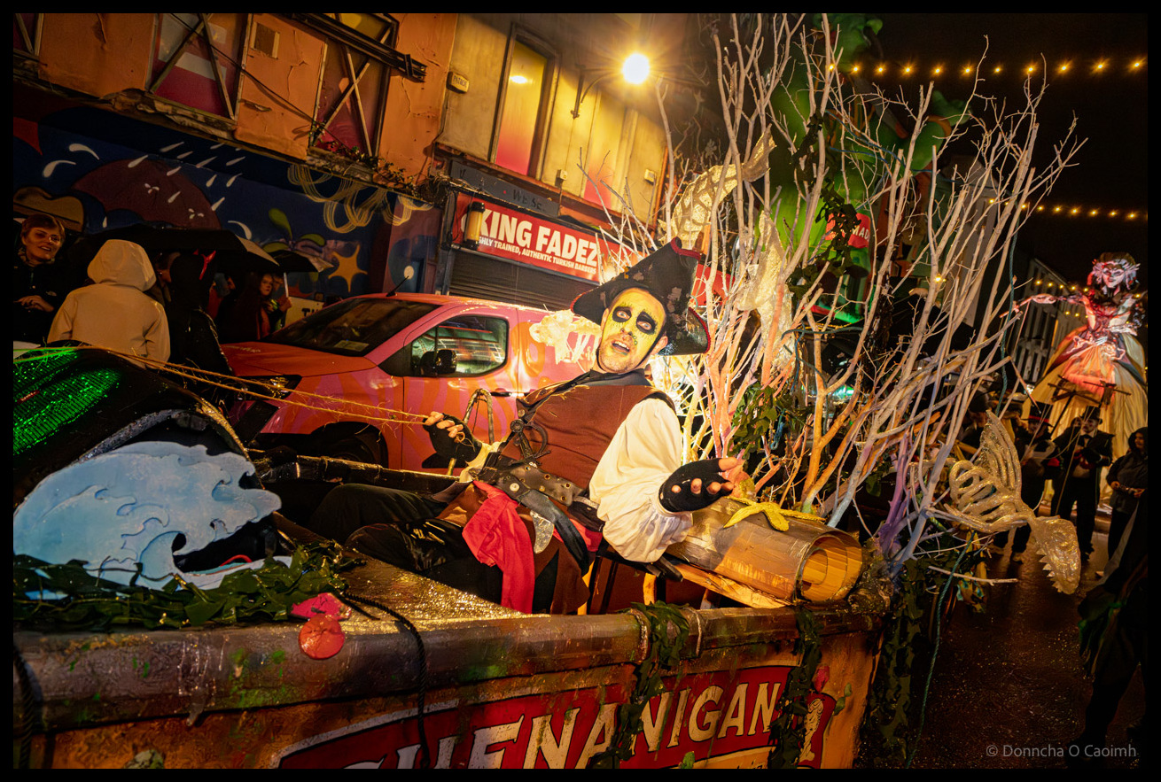 A participant in a pirate costume with skeleton face paint sits in decorated boat float labeled "SIENA VIGANO" surrounded by bare tree branches and theatrical props during Dragon of Shandon parade on North Main Street, Cork, with King Fadez shop and colourful murals behind.