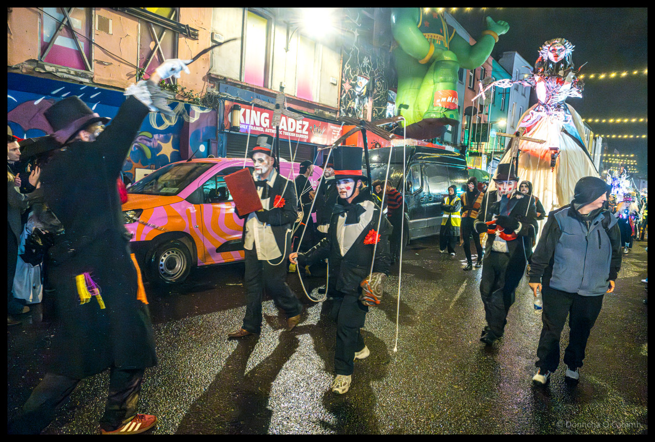 Wide view of Dragon of Shandon parade on North Main Street, Cork, showing participants in Victorian costumes manipulating puppets on strings, pink and orange striped van, large green dragon decoration overhead, elaborate tall female puppet figure on right, and King Fadez shop with colourful murals in background under string lights at night.