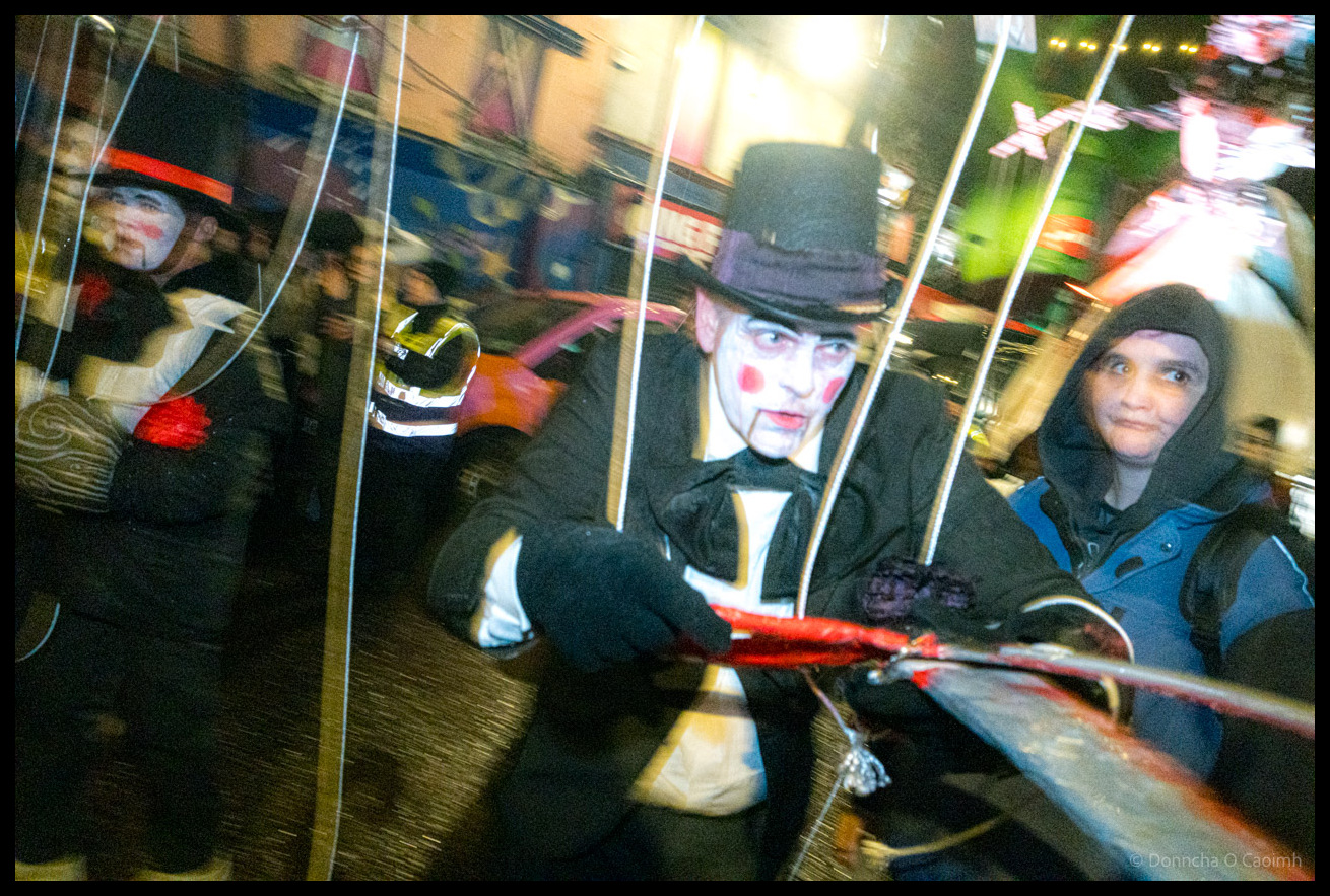 Motion-blurred close-up photograph of Dragon of Shandon parade participant in top hat and theatrical vampire-style makeup manipulating marionette strings with spectator visible on right on North Main Street, Cork, at night.