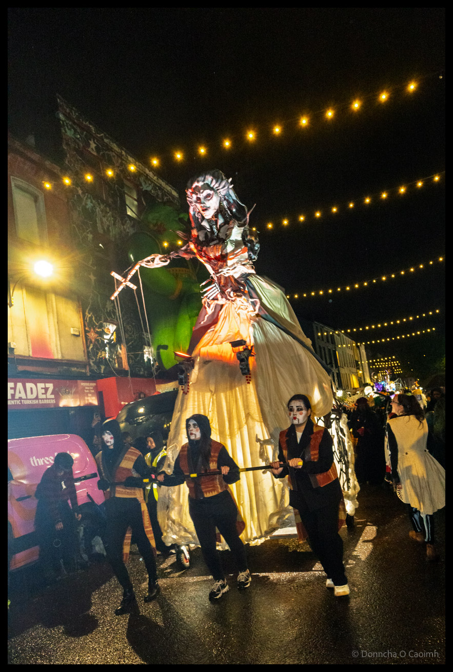 Tall elaborate female puppet figure in golden dress with ornate theatrical makeup and headdress being manipulated by performers in black with burgundy sashes during Dragon of Shandon parade on North Main Street, Cork, under string lights at night, with the King Fadez shop visible in background.