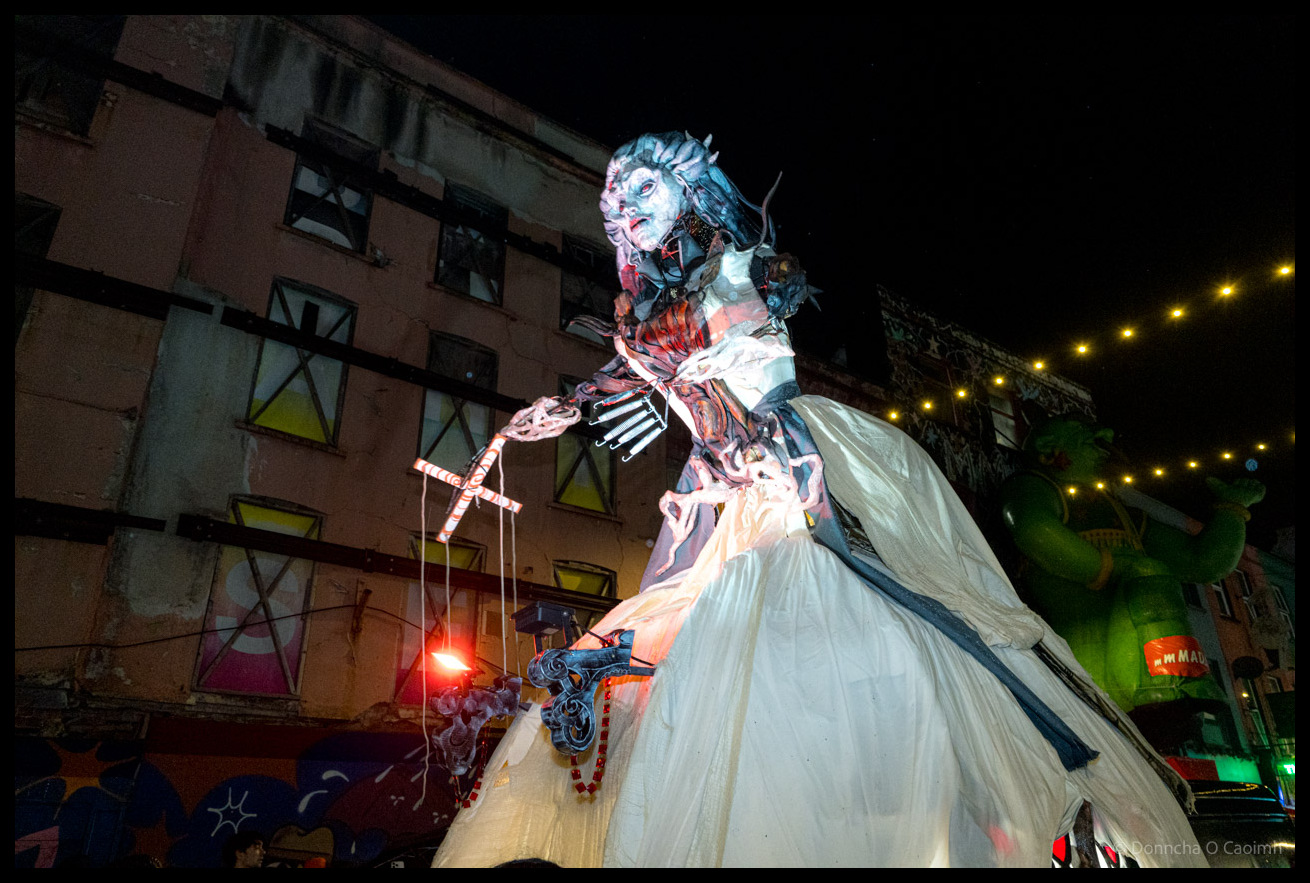 Close-up of towering female puppet figure with elaborate theatrical makeup, ornate costume details including skeletal hand design and flowing golden dress, during the Dragon of Shandon parade on North Main Street, Cork, at night with building windows visible behind.