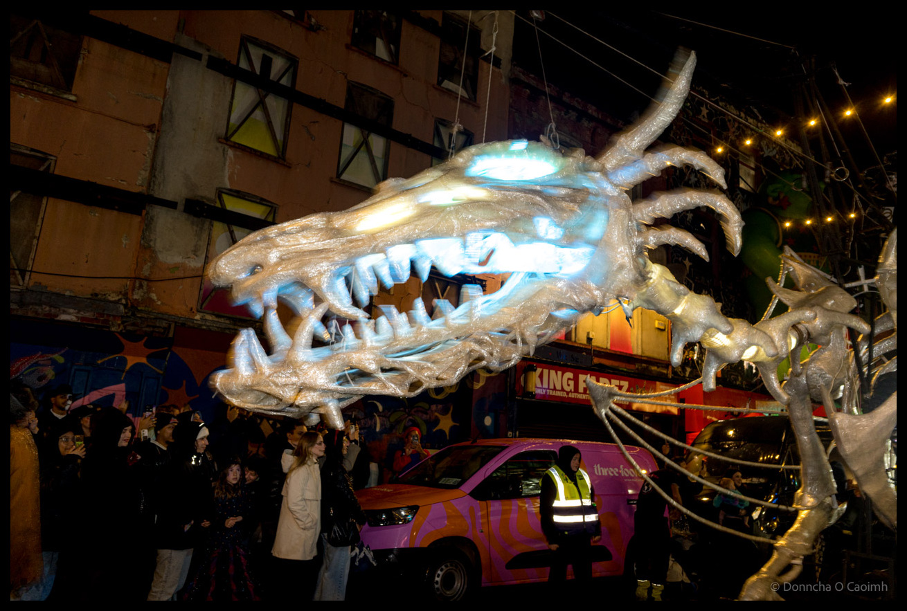 A massive illuminated dragon skull head with glowing blue interior and silver-painted skeletal texture towers over North Main Street, Cork, during the Dragon of Shandon parade at night, with a pink and orange striped van, King Fadez shop, colourful murals and crowds visible below.