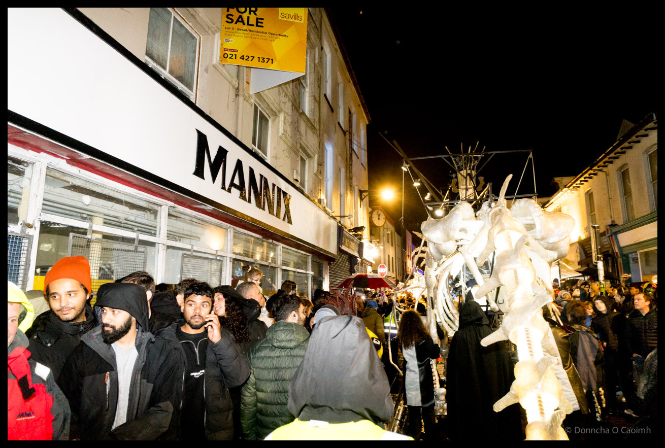 Crowds line Castle Street Cork outside Mannix shop watching massive white skeletal dragon puppet pass during Dragon of Shandon parade at night with spectators filming on phones