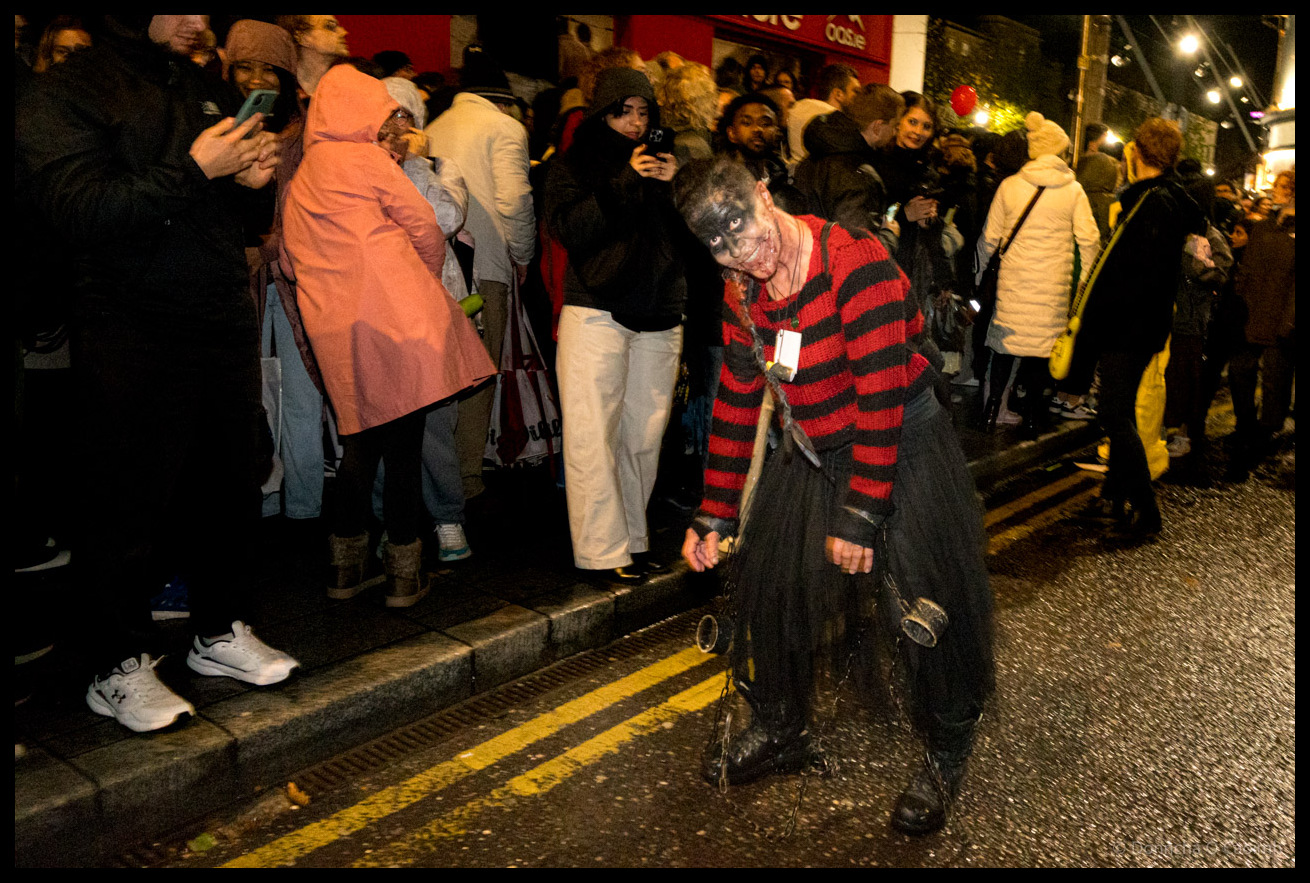 Young person in red and black striped costume with zombie makeup and holding knife poses dramatically in foreground whilst crowds of spectators line Cornmarket Street Cork watching Dragon of Shandon parade at night