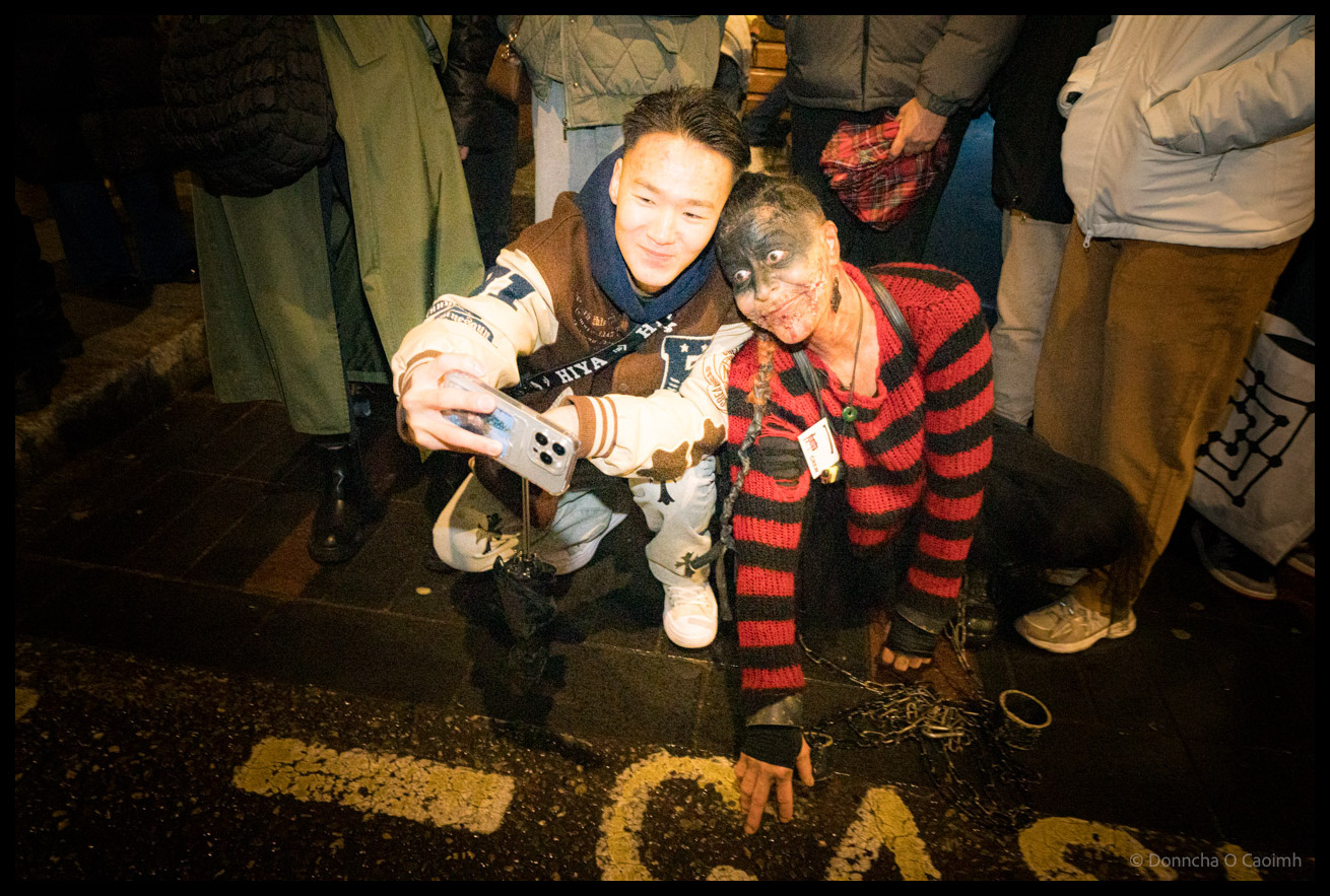 Spectator crouches to take selfie with parade participant in red and black striped zombie costume holding plastic knife on wet Cornmarket Street Cork pavement during Dragon of Shandon parade at night with crowds visible behind