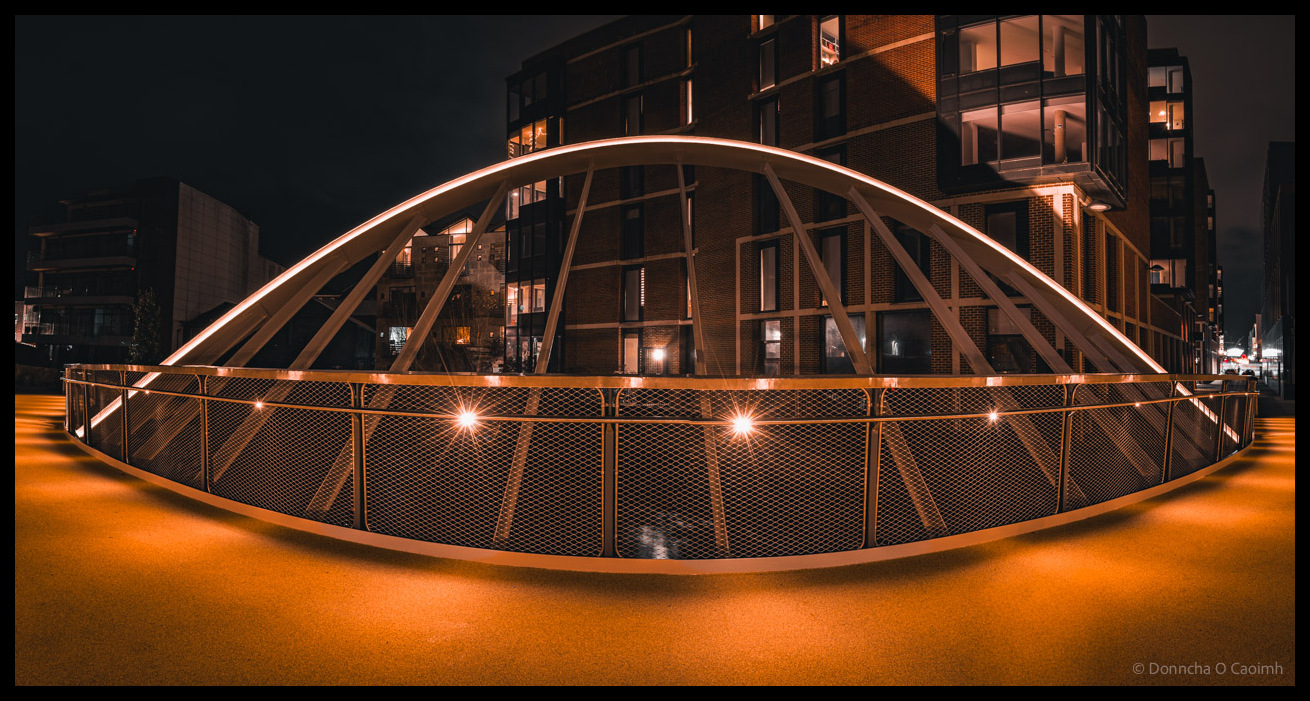 Modern curved pedestrian and cycle bridge with illuminated safety mesh fencing and architectural metal arch spanning the river from Lambley’s Lane (off South Main Street) to Wandesford Quay at night, with contemporary apartment buildings visible in background and orange-lit pathway surface creating dramatic urban nightscape.