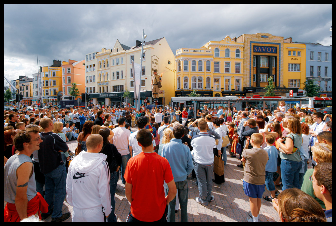 Photograph of a large crowd gathered on St. Patrick Street in Cork city centre in June 2006, watching a street performer on stilts (a fire breather) elevated above the crowd. The scene shows hundreds of spectators forming a circle around the performer, with people sitting and standing on the wide pavement. Visible storefronts include the yellow Savoy building with "Quills EST 1939" signage, Dunnes Stores with its green front, and colourful Georgian buildings in yellow, orange, cream and white. A Securicor vehicle and city bus are visible in the background. Dramatic cloudy sky overhead contrasts with sunlight illuminating the crowd. People wear casual summer clothing including a white Nike Air hoodie and FC Barcelona "Ronaldinho" jersey.