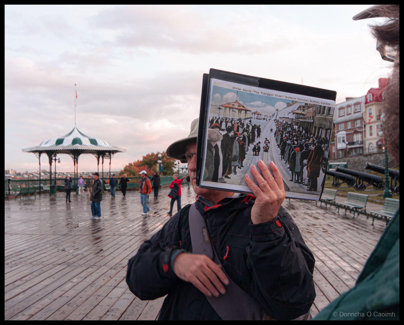 Photograph of a tour guide on Dufferin Terrace in Quebec City, Canada, holding up a framed historical image titled "Winter Sports 'The Toboggan Slide' Dufferin Terrace, Quebec, Canada" showing crowds of people in Victorian-era clothing watching tobogganing. The guide wears a wide-brimmed hat, glasses, and a black jacket with red trim, with a bag strap visible. The wooden boardwalk is wet from rain, with tourists walking in the background near a distinctive green and white striped gazebo bandstand. Autumn foliage, historic cannons, green benches, and traditional Quebec City buildings are visible. A Canadian flag flies in the distance under an overcast pink-tinged evening sky.