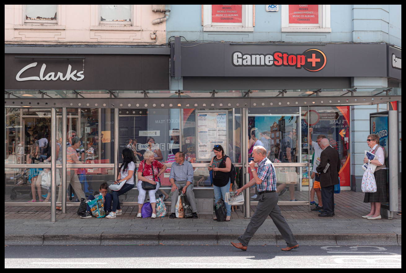 Street photography scene showing a busy bus shelter outside Clarks and GameStop+ storefronts with multiple passengers waiting and standing, including children, elderly people, and various pedestrians in casual summer clothing, Victorian-era building with pink and blue facade visible above, red shop signage, daytime urban street setting, taken July 2017.