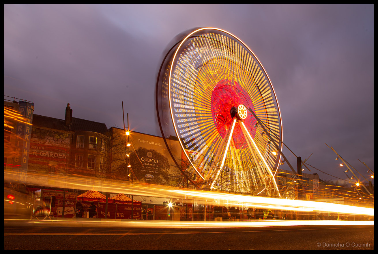 Long exposure photograph of the Cork Christmas Ferris wheel on Grand Parade at dusk in December 2017. The wheel displays a striking pattern with a red illuminated centre, yellow and orange spokes radiating outward, and a white rim created by the spinning lights. Golden vehicle light trails streak across the foreground. A purple-blue twilight sky forms the backdrop. Visible signage includes "PARKING", "BEER GARDEN", and a billboard reading "Talent will out..." advertising "QUERCUS Scholarship" for "University College Cork" with a Ford logo. Red and white striped Christmas market stalls sit at the base of the wheel.