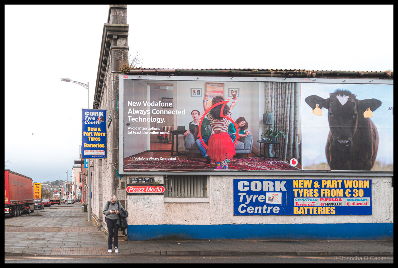 Street photograph of a woman in grey hoodie and black leggings looking at her phone while standing on a pavement in Cork, Ireland. Above her is a large Vodafone billboard reading "New Vodafone Always Connected Technology. Avoid interruptions (at least the online ones)." with a search bar showing "Vodafone Always Connected" and featuring a family scene in a living room. Adjacent billboard shows two black cattle with yellow ear tags and text "together we can" with Vodafone logo. Below are signs for "Cork Tyre Centre" advertising "NEW & PART WORN TYRES FROM €30" and "BATTERIES" with logos for Goodyear, Fulda, Pirelli, Michelin, Hankook, and Continental. A "Pzazz Media" sign and bilingual street sign reading "SRÁID NA LONG / SHIP STREET 1" are visible. Left side shows another Cork Tyre Centre sign with "New & Part Worn Tyres Batteries Tel: 083 1980602". Urban street with parked vehicles and lorries extends into the distance.