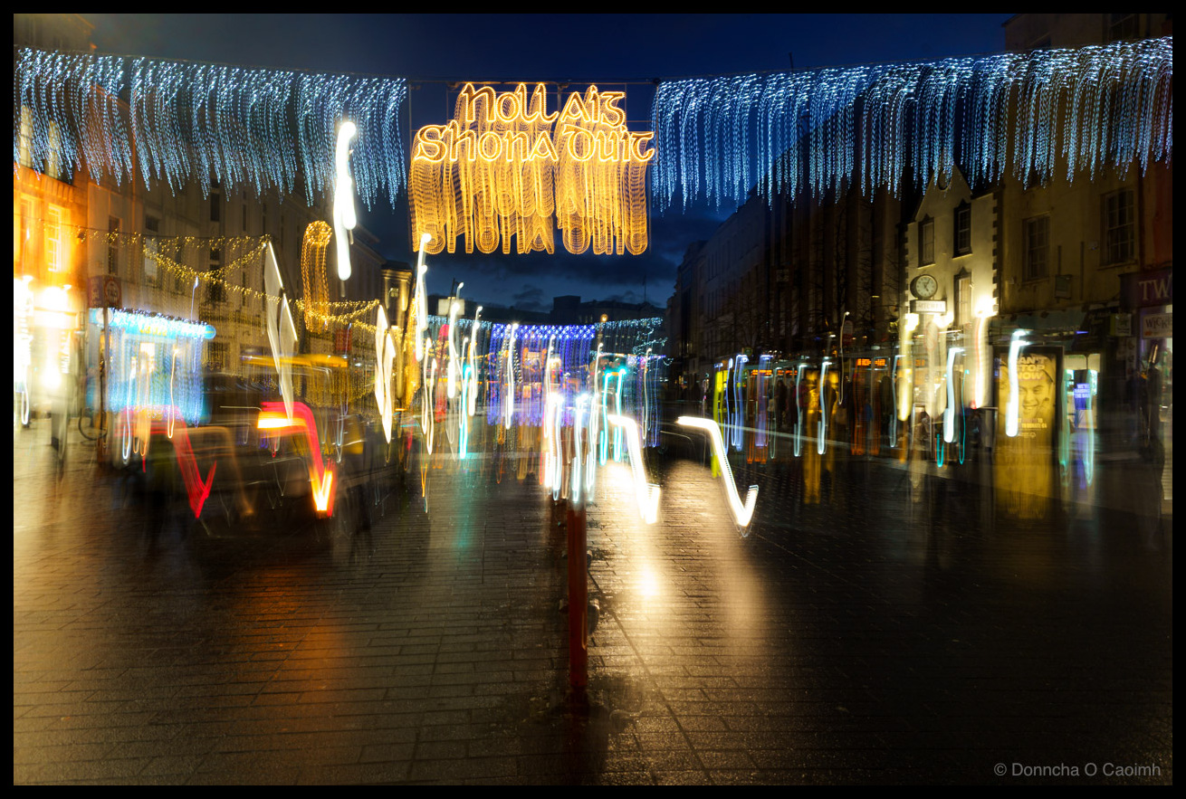 Abstract long-exposure photograph of Christmas decorations on St. Patrick Street, Cork in December 2022. The image features intentional camera movement creating streaks and blur throughout. The sharp focal point is the illuminated Irish language sign reading "Nollaig Shona Duit" (Happy Christmas to you) in warm golden yellow lights hanging above the street. Blue icicle-style LED lights cascade down from above like a frozen waterfall. The motion blur transforms shopfronts, street lights and pedestrians into colourful streaks of light in red, yellow, white and blue. The wet pavement reflects the lights creating mirror effects. Buildings line both sides of the street under a dark evening sky.