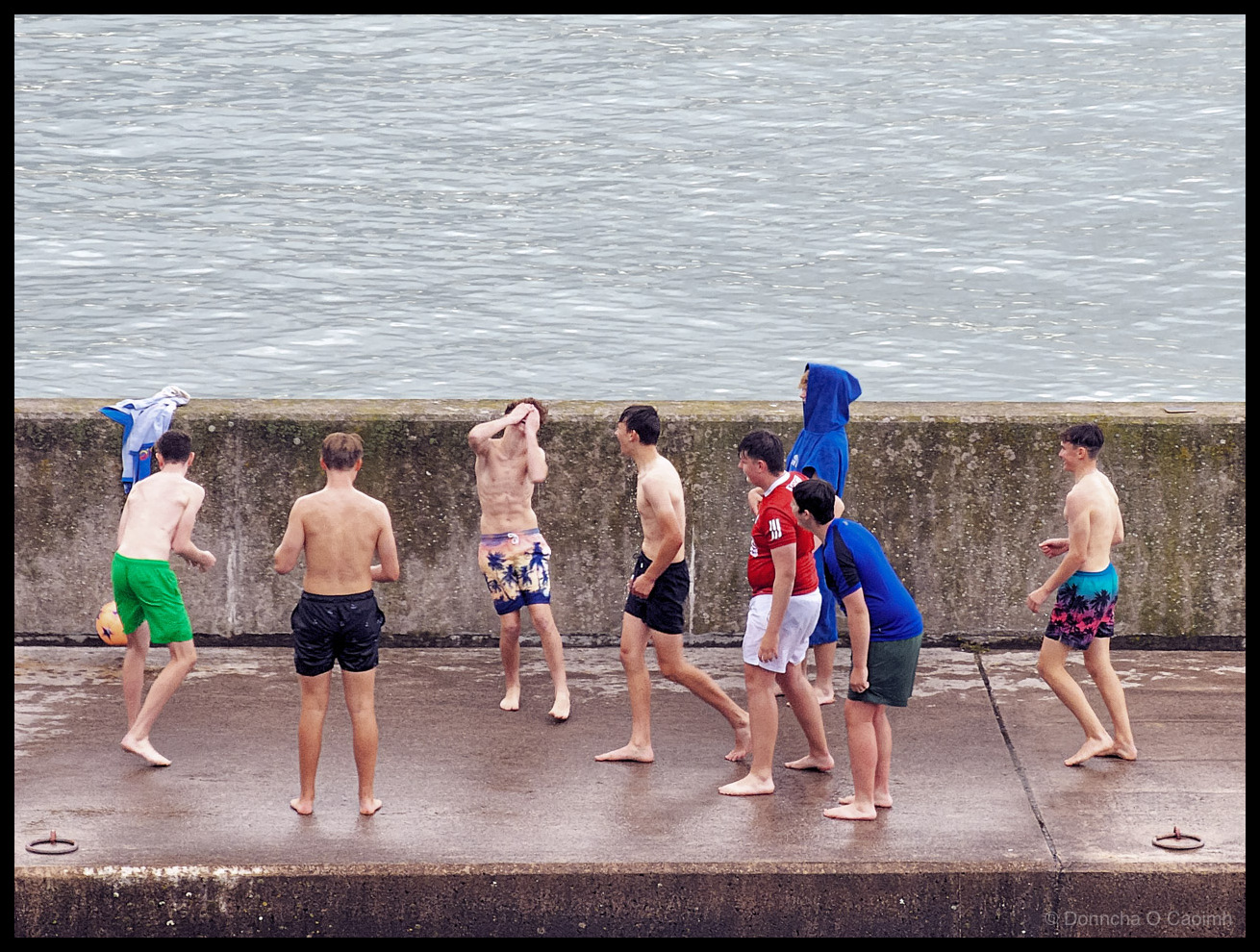 Photograph of a group of eight boys and teenagers on the wet concrete slipway at Ardmore Boat Cove, County Waterford, Ireland. Most are shirtless and barefoot in colourful swim shorts including green, black, blue with palm tree print, and black with pink palm trees. A football lies on the ground behind them, one boy has a blue T-shirt on, one wears a red Manchester United jersey, and another wears a blue hoodie with the hood up. They stand along the concrete pier wall with calm grey sea water behind them. Metal mooring rings are visible on the wet concrete surface.