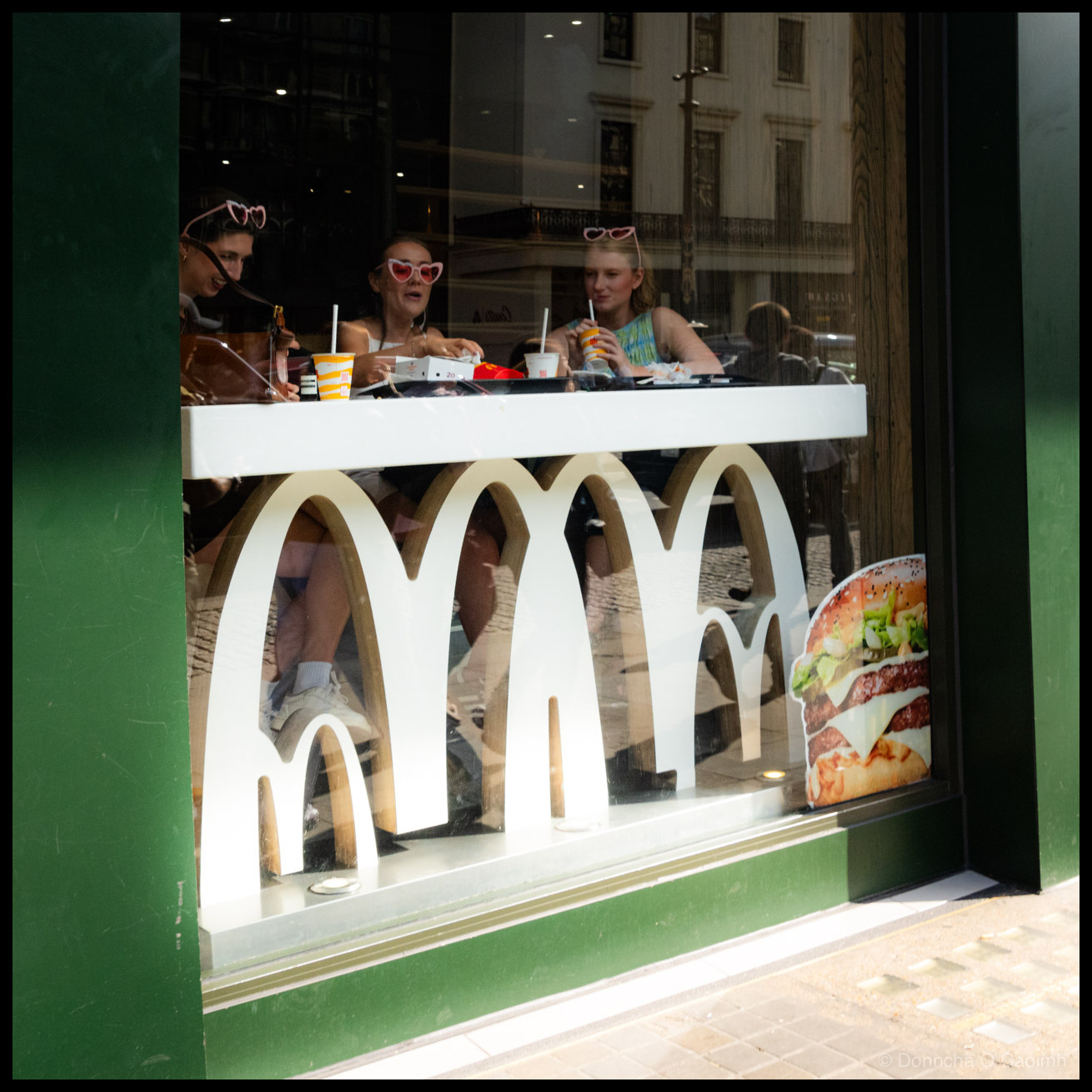 Street photograph looking through the window of a McDonald's restaurant in London, UK, during summer 2025. Three women wearing matching pink heart-shaped sunglasses sit at a window counter eating and drinking. One wears a grey top, one a white strappy top, and one a colourful patterned top. McDonald's cups with yellow striped designs and food packaging sit on the white counter in front of them. The white McDonald's golden arches logo on the window frames them from below, while a large burger advertisement is visible in the lower right corner. Street reflections including pedestrians and a classical building facade are visible in the glass. The restaurant exterior is painted dark green. 