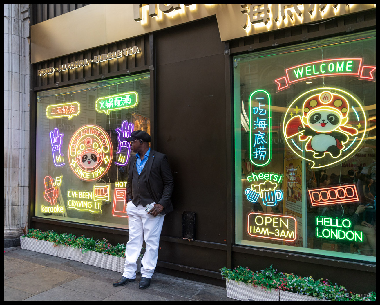 Street photograph of a well-dressed man standing outside a Haidilao Hot Pot restaurant in London, UK in summer 2025. He wears a dark grey blazer, light blue open-collar shirt, white trousers, black shoes and a black flat cap, leaning casually against the dark brown storefront while holding a phone. The restaurant windows are covered in colourful neon signs including a panda mascot with "WELCOME" banner, "HELLO LONDON", "OPEN 11AM-3AM", "cheers!" with beer mugs, "karaoke", "I'VE BEEN CRAVING IT", "HOT", "HAIDILAO HOT POT SINCE 1994" with panda logo, and Chinese characters including "三五好友" (a few good friends), "火锅配酒" (hot pot with alcohol), and "一吃海底捞" (eat Haidilao). Above the windows text reads "FOOD • ALCOHOL • BUBBLE TEA". Flower planters sit beneath the windows.