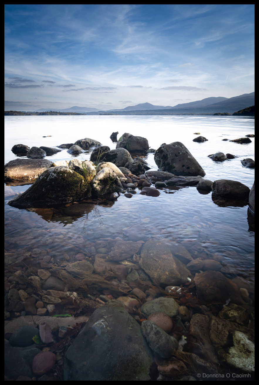 A photograph of the Lakes of Killarney near O'Sullivan's Cascade showing a rocky foreshore with moss-covered boulders in crystal-clear shallow water, mountains and forested shoreline visible across the calm lake under blue sky with white clouds, taken October 2025.