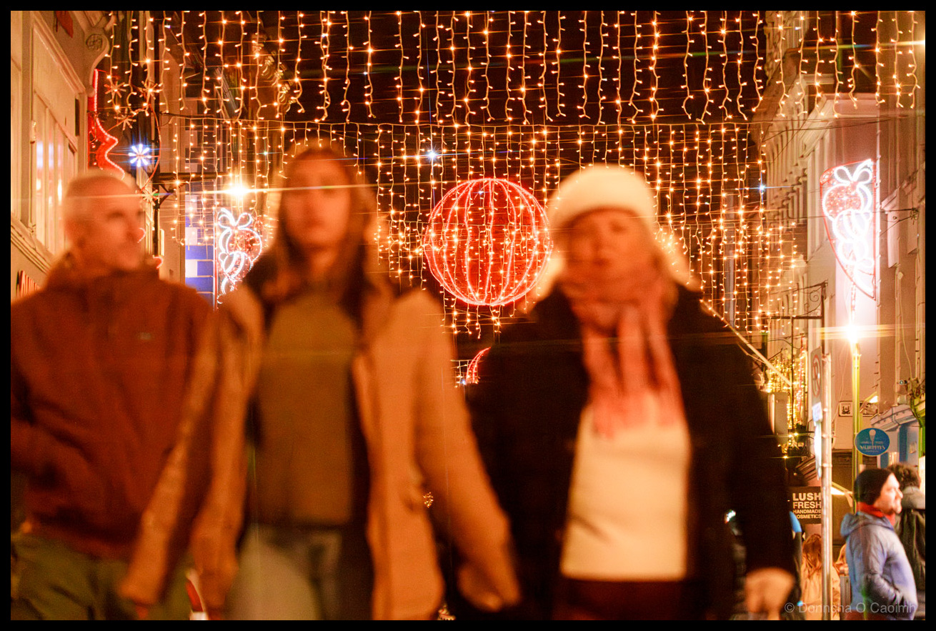 Night-time photograph of pedestrians walking on Oliver Plunkett Street, Cork, beneath cascading golden fairy lights creating a canopy effect overhead, large red neon-illuminated festive spheres and decorative motifs suspended above the street, blurred figures in winter clothing in motion, storefront signage visible at street level.