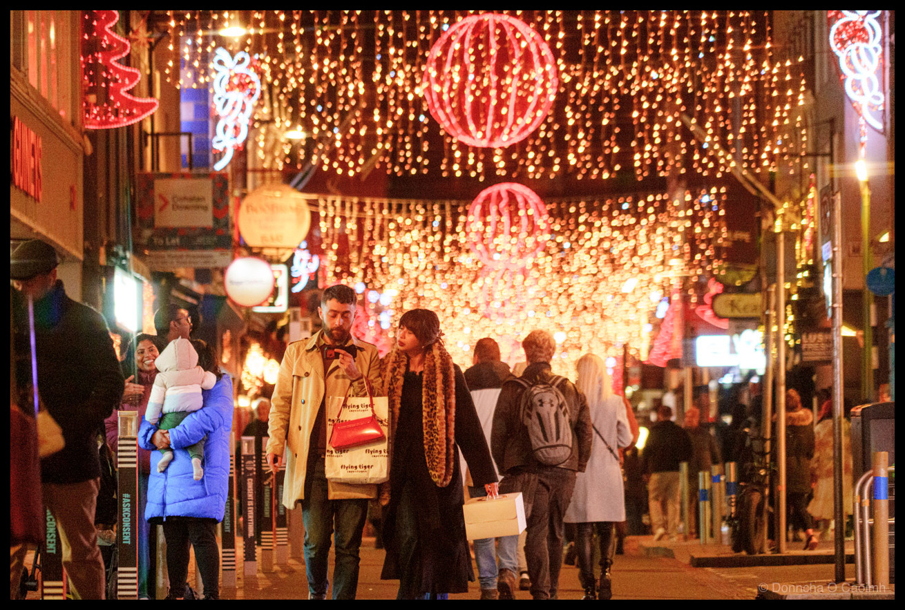 Night-time photograph of pedestrians walking on Oliver Plunkett Street, Cork, beneath cascading golden fairy lights creating a canopy effect overhead, large red neon-illuminated festive spheres and decorative motifs suspended above the street, blurred figures in winter clothing in motion, storefront signage visible at street level.