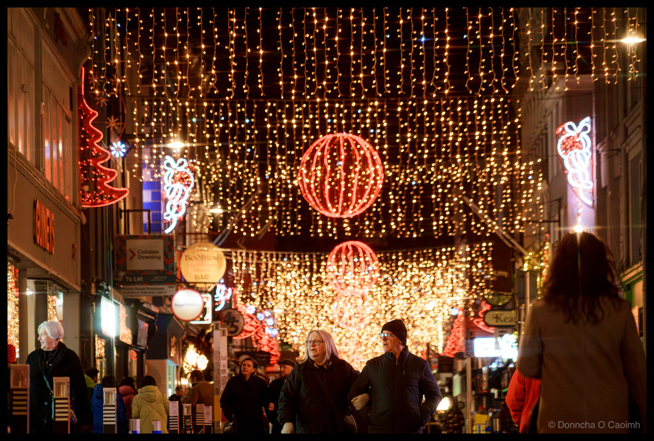 Night-time photograph of pedestrians walking on Oliver Plunkett Street, Cork, beneath cascading golden fairy lights creating a canopy effect overhead, large red neon-illuminated festive spheres and decorative motifs suspended above the street, blurred figures in winter clothing in motion, storefront signage visible at street level.
