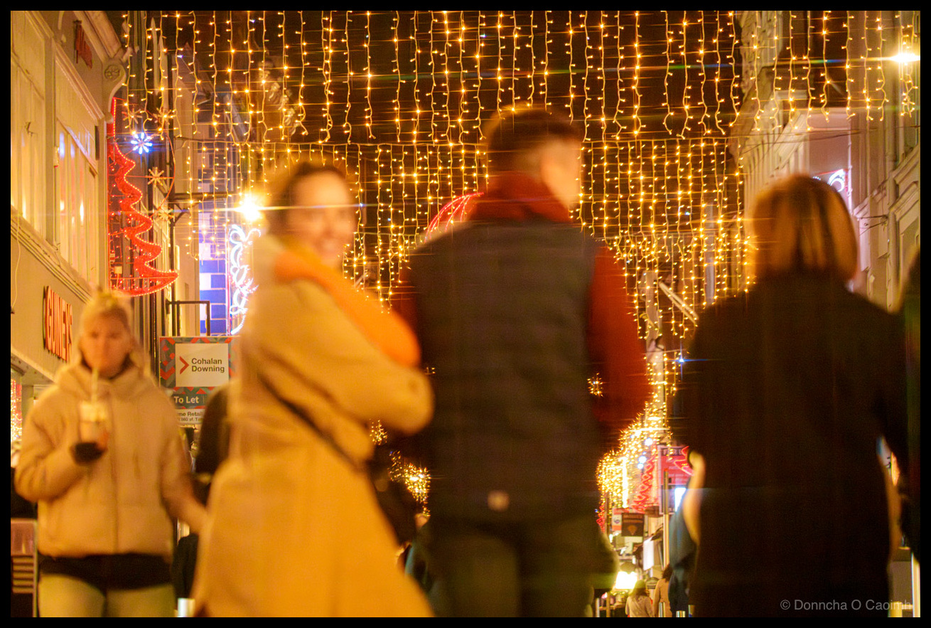 Night-time photograph of pedestrians walking on Oliver Plunkett Street, Cork, beneath cascading golden fairy lights creating a canopy effect overhead, large red neon-illuminated festive spheres and decorative motifs suspended above the street, blurred figures in winter clothing in motion, storefront signage visible at street level.