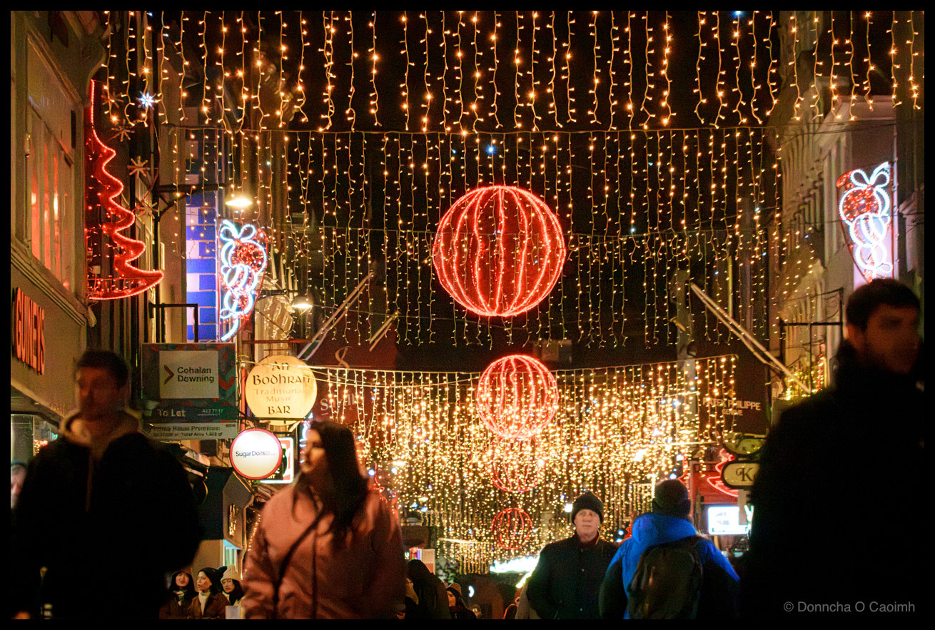 Night-time photograph of pedestrians walking on Oliver Plunkett Street, Cork, beneath cascading golden fairy lights creating a canopy effect overhead, large red neon-illuminated festive spheres and decorative motifs suspended above the street, blurred figures in winter clothing in motion, storefront signage visible at street level.