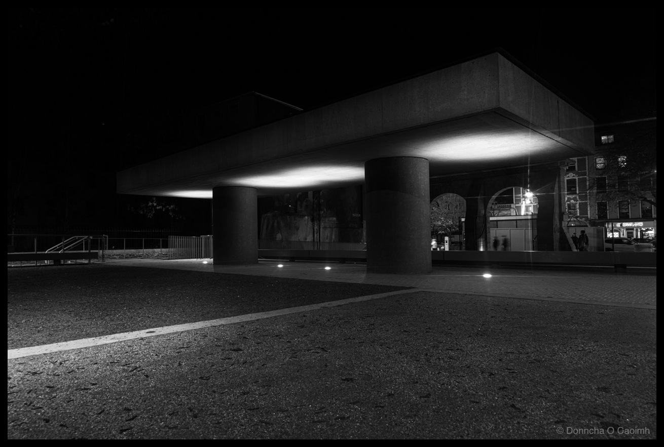 Black and white night-time photograph of a modern brutalist covered structure with horizontal fluorescent strip lighting and concrete cylindrical support pillars, gravel and paved ground in foreground, white metal railings, modern building visible on right side, and a contemporary public space at Bishop Lucey Park, Cork.