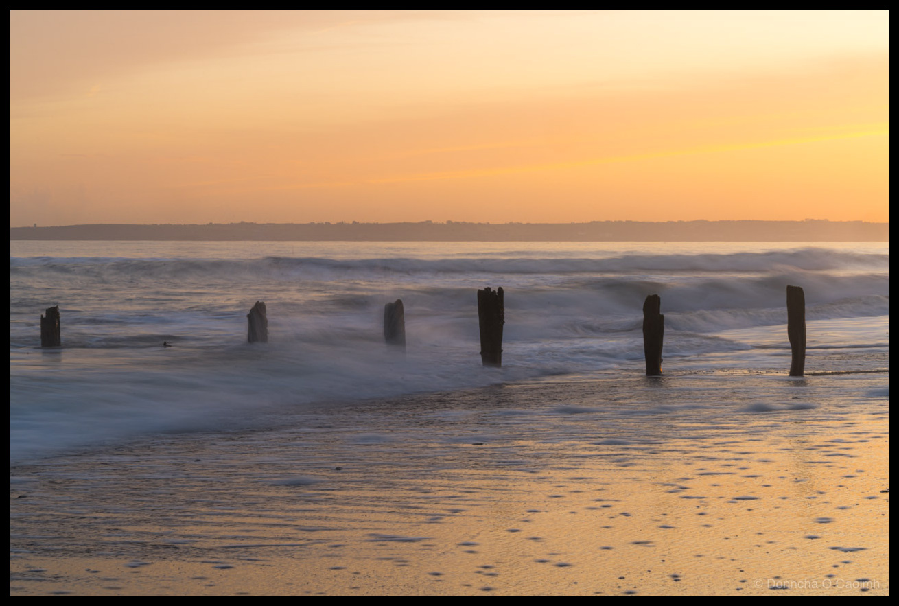 Long exposure sunset photograph of Youghal beach, County Cork, Ireland in December 2025. Weathered wooden posts, remnants of old groynes stand silhouetted in the incoming surf. The long exposure has rendered the waves as soft, misty swirls of white and blue water around the dark timber posts. The sky glows in warm tones of peach, orange and soft yellow, with a distant headland visible on the horizon. The wet sand in the foreground reflects the golden sunset light, with small pools and ripples catching the colour.