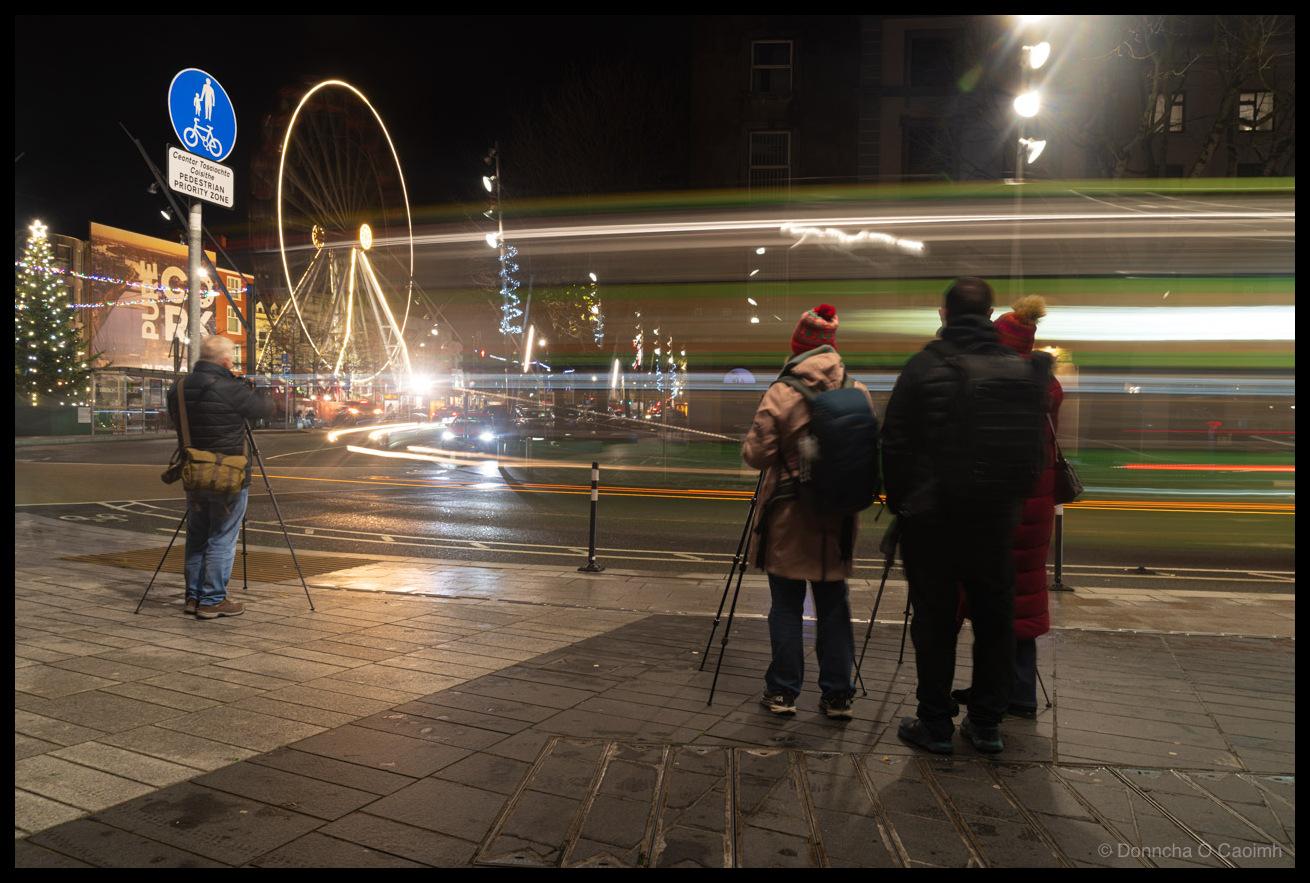 Long exposure night photograph showing a group of photographers with tripods spread along Grand Parade, Cork in December 2025. A bilingual "Ceantar Tosaíochta Coisithe / PEDESTRIAN PRIORITY ZONE" sign with bicycle and pedestrian symbol is visible on the left. Green and white light trails from a passing bus streak through the frame. The illuminated Cork Christmas ferris wheel and decorated Christmas trees are visible in the background near Bishop Lucey Park. One photographer stands alone on the left while a group of four cluster together on the right.