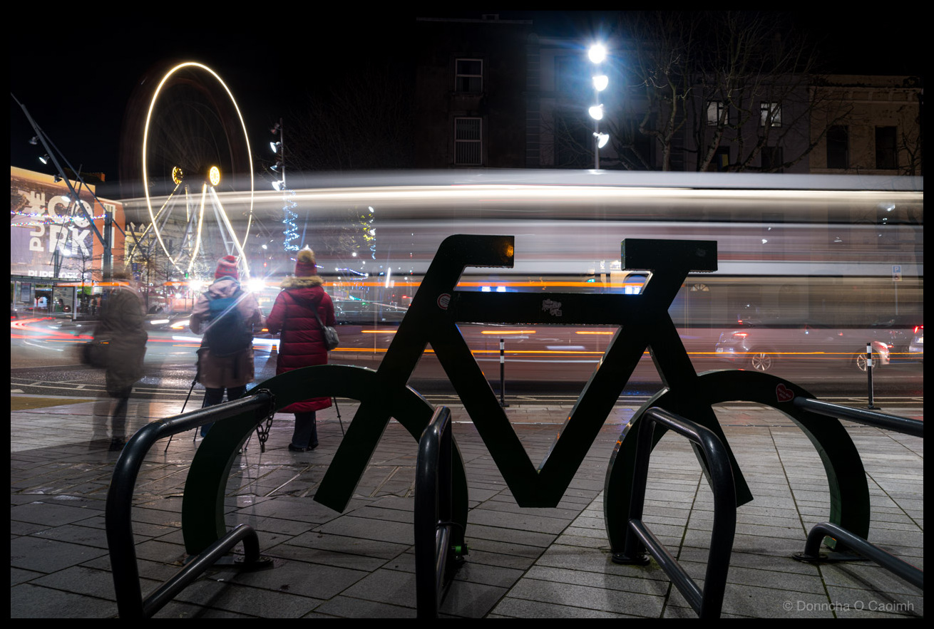 Long exposure night photograph featuring a large black bicycle-shaped bike rack sculpture in the foreground on Grand Parade, Cork in December 2025. Behind it, photographers with tripods are visible as slightly blurred figures, with a woman in a red puffer coat prominent among them. White bus light trails streak across the scene. The Cork Christmas ferris wheel is illuminated in the background, and Christmas tree lights are visible. The wet pavement reflects the city lights. 