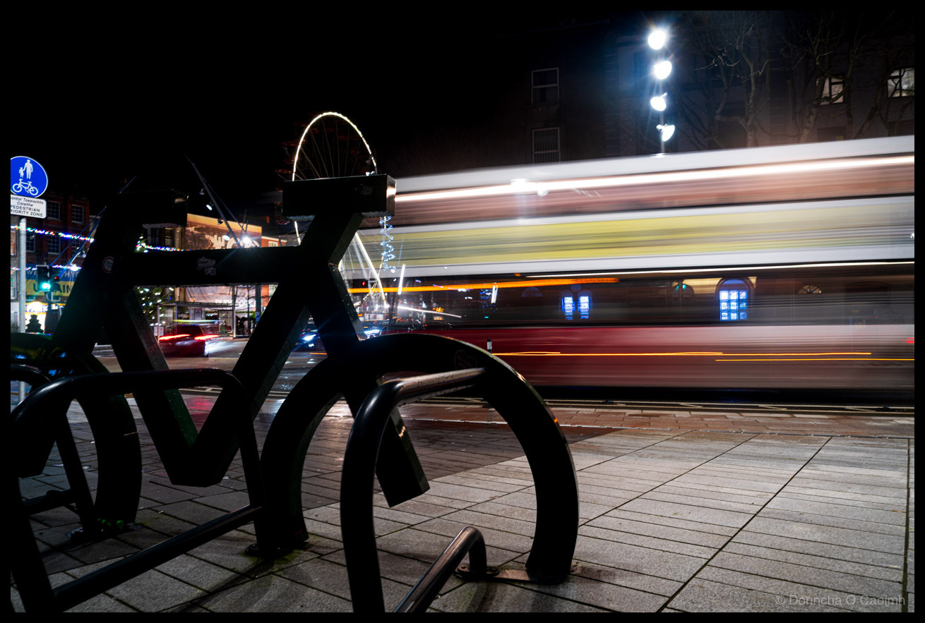 Long exposure night photograph of a black bicycle-shaped bike rack silhouetted against the lights of Grand Parade, Cork in December 2025. A Bus Éireann bus passes behind, creating horizontal red, cream and yellow light trails that reveal its livery through motion blur. The Cork Christmas ferris wheel is visible in the background. A blue pedestrian zone sign is partially visible on the left, and Christmas lights decorate buildings in the distance. Street lights create starburst effects in the dark sky.