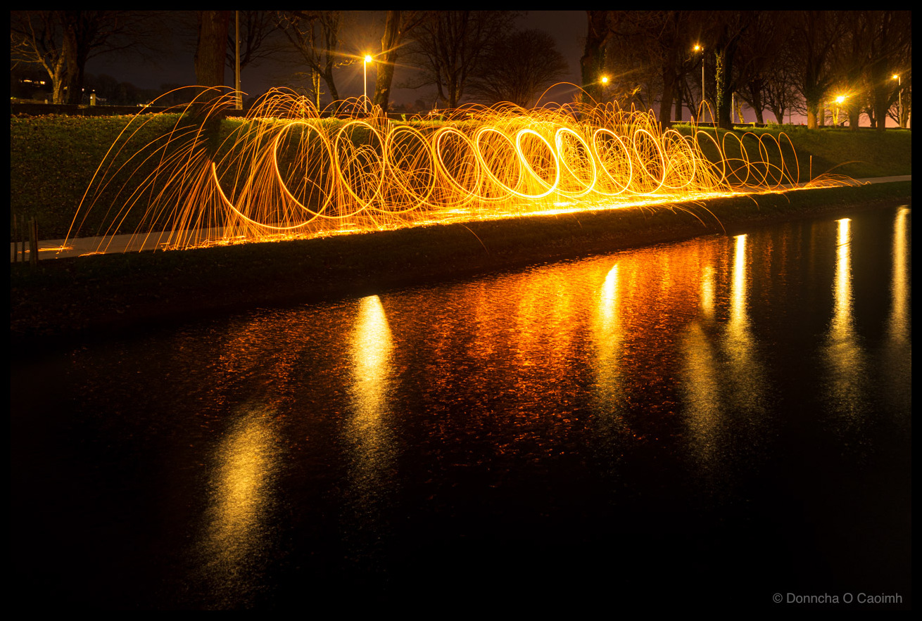 Long exposure photograph of steel wool spinning at night, taken in December 2025. Burning wire wool has been spun in circles while walking along a path beside a body of water, creating a continuous spiral of golden-orange sparks that forms a helix or spring-like pattern stretching across the frame. Individual spark trails arc outward from the main spiral. The sparks and their bright orange reflections shimmer in the dark water below, doubling the effect. Bare winter trees and street lights are visible in the background against a dark sky. A low hedge runs along the water's edge.