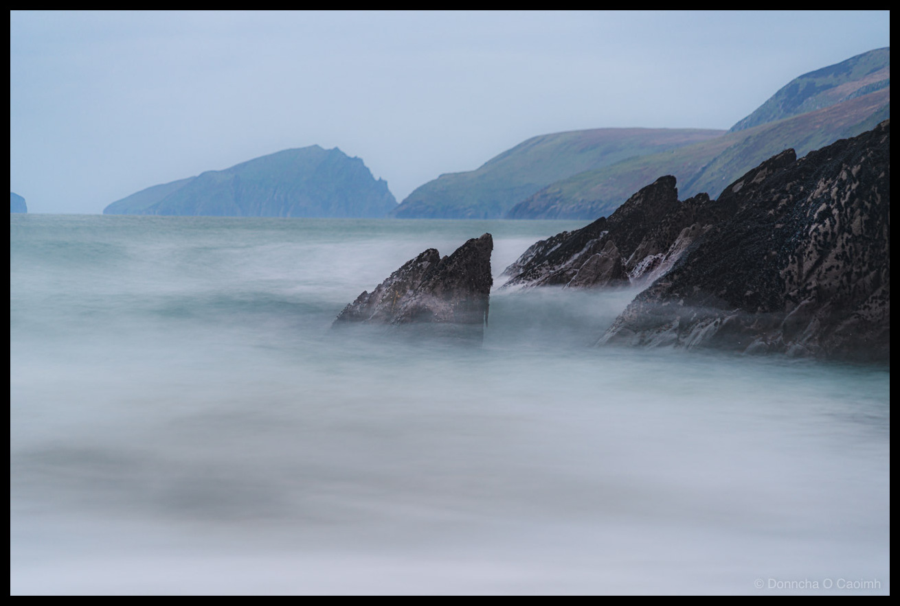 Long exposure landscape photograph of Couminole Beach on the Dingle Peninsula, County Kerry, Ireland in December 2025. Dark jagged rocks rise from the sea in the foreground, their rough barnacle-covered surfaces rendered sharp against the silky smooth water created by the extended exposure time. The Atlantic waves have been transformed into an ethereal white mist swirling around the base of the rocks. In the background, the dramatic headlands and mountains of the Dingle Peninsula recede into the grey overcast sky, their green and brown slopes fading into atmospheric haze. The colour palette is muted and moody in tones of grey, blue-green, and brown.