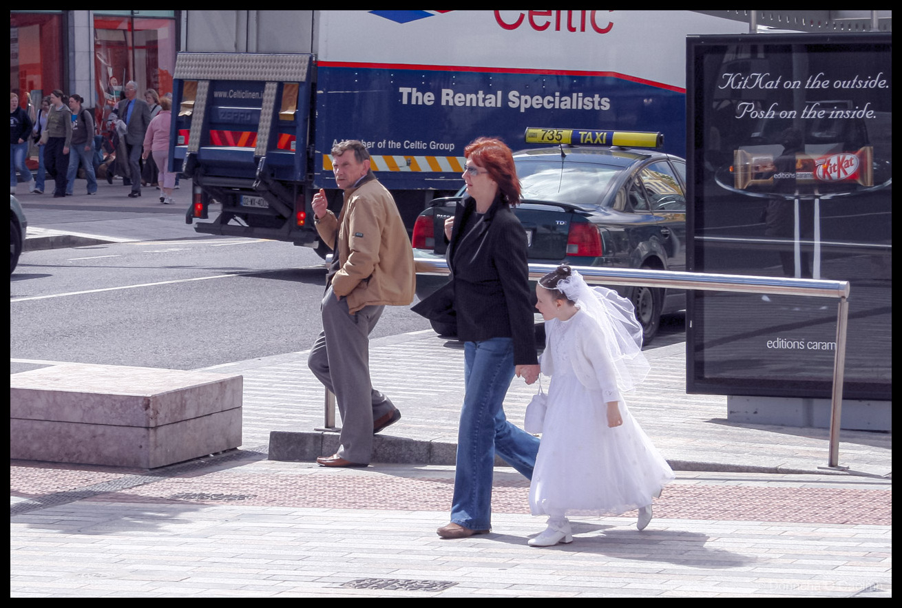 Street photograph on St. Patrick Street in Cork, Ireland showing a young girl in a white First Holy Communion dress with veil, tiara, and white handbag walking hand-in-hand with her mother who has red hair, glasses, and wears a black jacket with blue jeans. A man in a beige jacket and grey trousers leans against a barrier. Background shows a blue Celtic Linen truck with "The Rental Specialists" and "A Member of the Celtic Group" text, a black taxi with yellow "735 TAXI" sign, and shoppers on the street. On the right is a KitKat advertisement reading "KitKat on the outside. Posh on the inside." with "editions caramel" below.