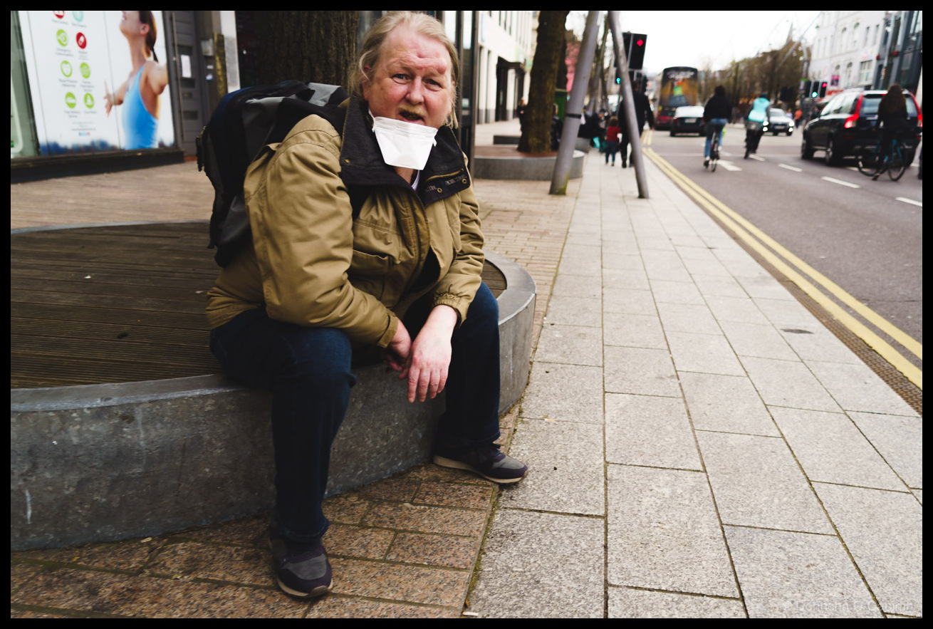 Street photography showing a man wearing a white face mask and tan jacket with black backpack, sitting on a concrete street planter on a busy urban pavement in Cork, Ireland during the COVID-19 pandemic.