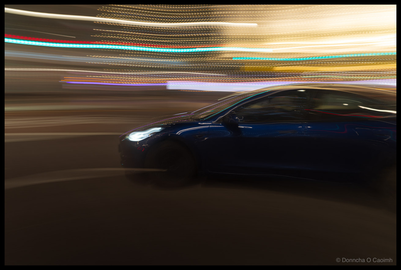 Long exposure panning photograph of a dark blue Tesla driving at night with colourful light trails streaking across the background in cyan, red, orange and yellow, creating a sense of speed and motion.