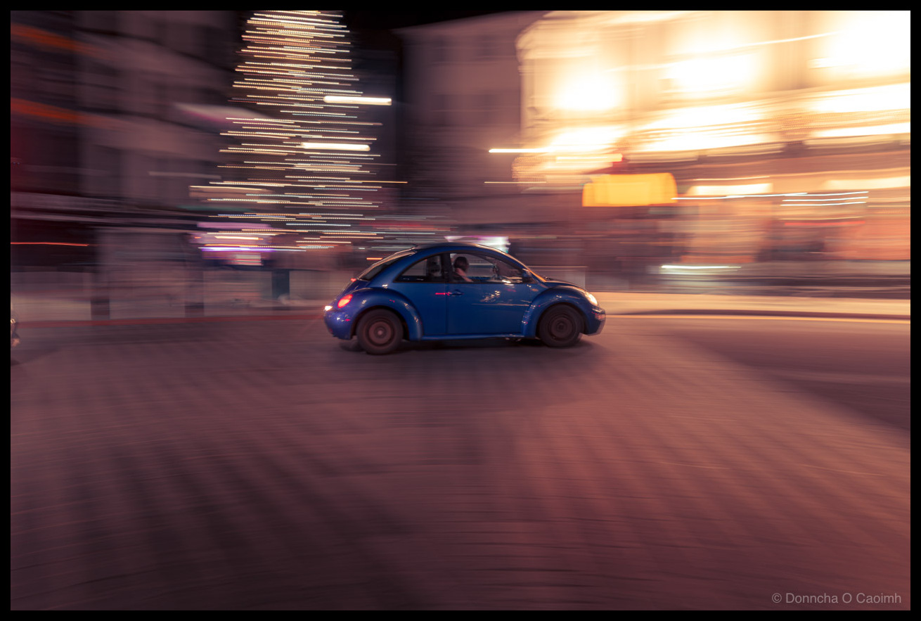 Motion blur panning shot of a blue Volkswagen New Beetle driving through a city at night, with a blurred Christmas tree light display and warm glowing shopfronts in the background.