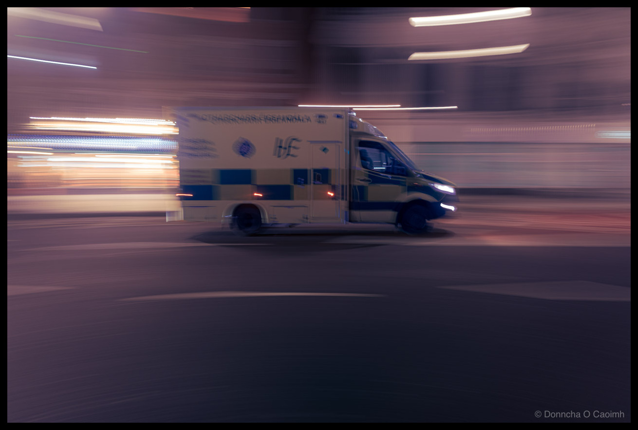 Panning photograph of an Irish HSE ambulance speeding through the city at night, showing yellow and blue Battenberg markings and text reading OTHARCHARR ÉIGEANDÁLA and HSE, with streaked urban lights in the background.