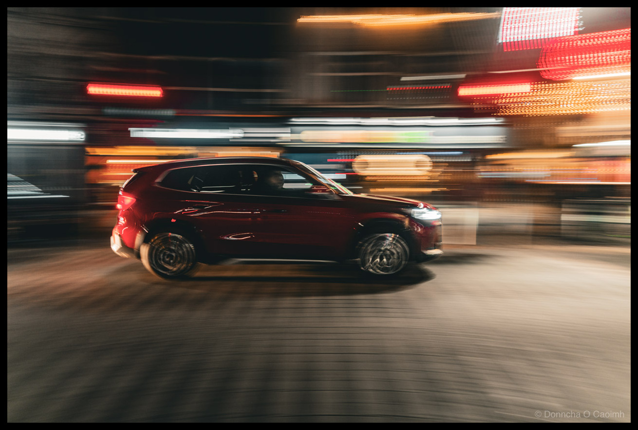 Night-time panning shot of a dark red SUV driving through a city street, with motion-blurred neon signs and colourful urban lights creating horizontal streaks in the background.