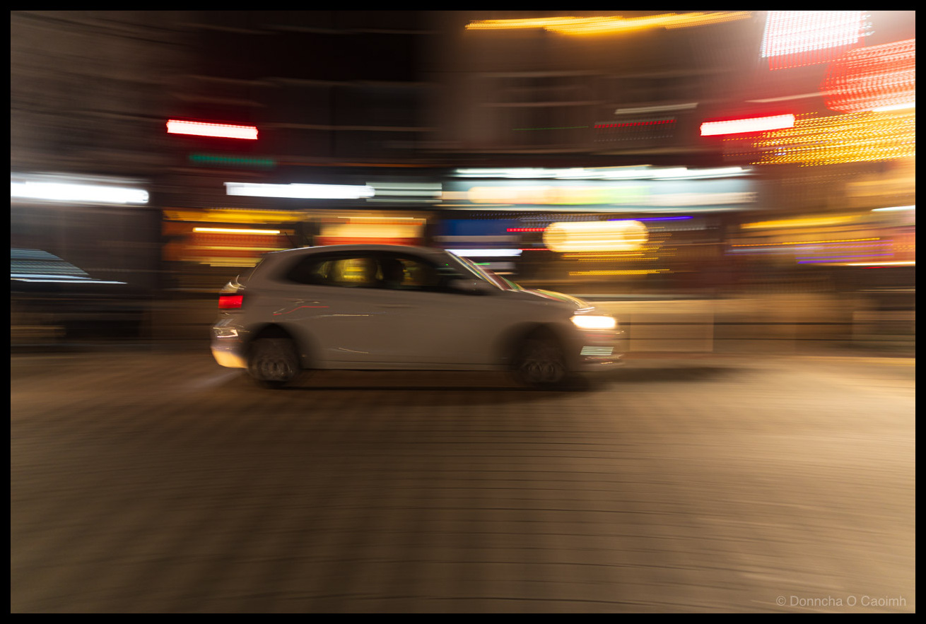 Motion blur photograph of a white hatchback car driving at night, captured using panning technique with vibrant bokeh and light trails from city signs and shopfronts blurred in the background.