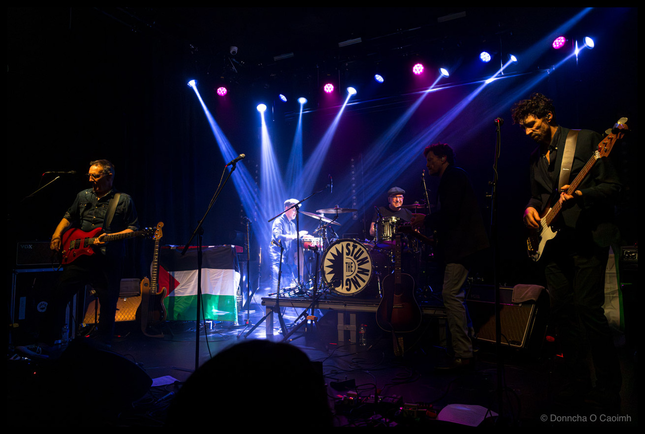 Concert photograph of Irish band The Stunning performing on stage at Cyprus Avenue, Cork, with blue and magenta spotlights cutting through the darkness, featuring four band members including a guitarist with a red electric guitar, lead singer in a white hat and pale jacket, drummer behind a kit displaying The Stunning logo, and bassist on the right, with a Palestinian flag visible on stage.