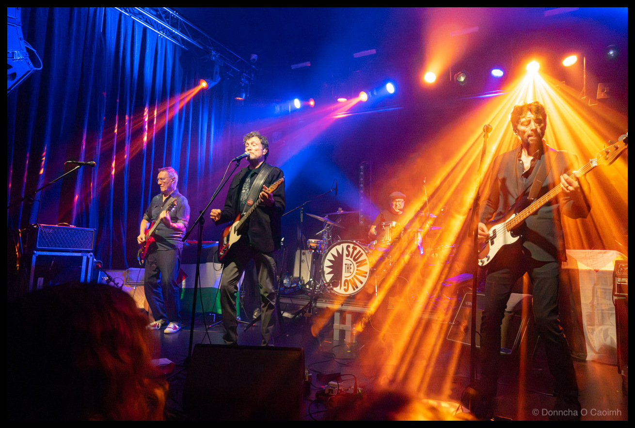 The Stunning performing live with dramatic orange and blue stage lighting, showing the lead singer centre stage playing guitar and singing, a guitarist with a red electric guitar on the left, the bassist bathed in golden light on the right, and the drummer visible behind The Stunning branded bass drum, with audience silhouettes in the foreground.