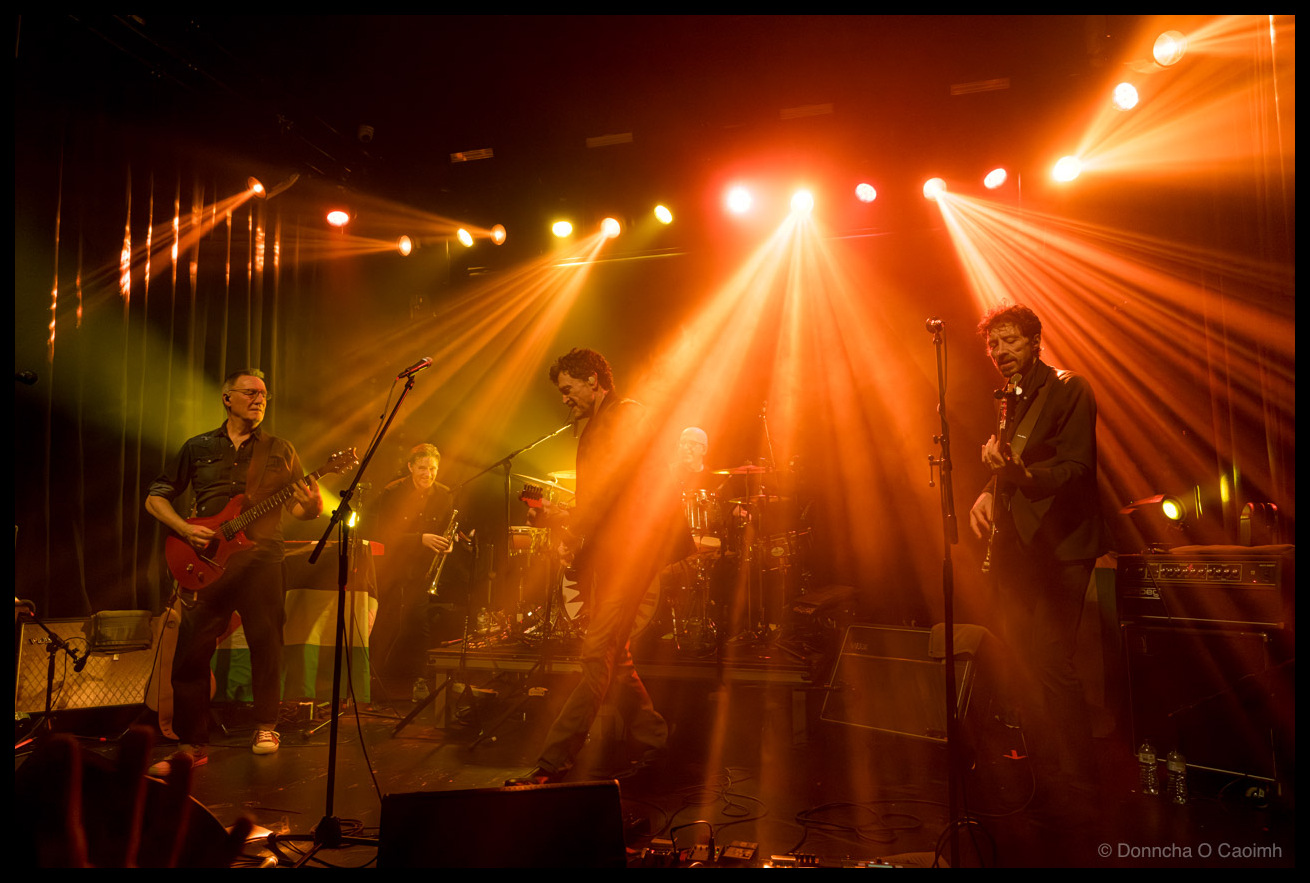 Wide stage shot of The Stunning performing with warm orange and yellow lighting beams, featuring five musicians on stage including a guitarist with a red electric guitar on the left, a brass player, the drummer, the bassist walking across stage, and another guitarist on the right.
